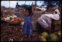 A young girl stands near a pile of cassava in a market with a bowl of fruit atop her head, Managua, Nicaragua