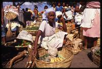 An elderly woman sits among baskets of plantains, peppers, and cassava at Eastern Market, Managua, Nicaragua