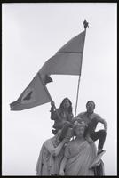 An alternate view of two young men holding a United Farm Workers of America flag and a National Liberation Front (Viet Cong) flag while sitting atop the Peace monument during the Vietnam War Out Now demonstration, 24 April 1971