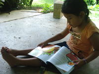 A young girl sits on a concrete porch reading a children's book while wearing Rachel Teter's glasses, El Plátano, Panama