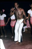 Man with microphone in white pants performing with dancers and band