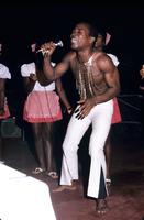 Man in white pants singing into microphone in front of three female dancers